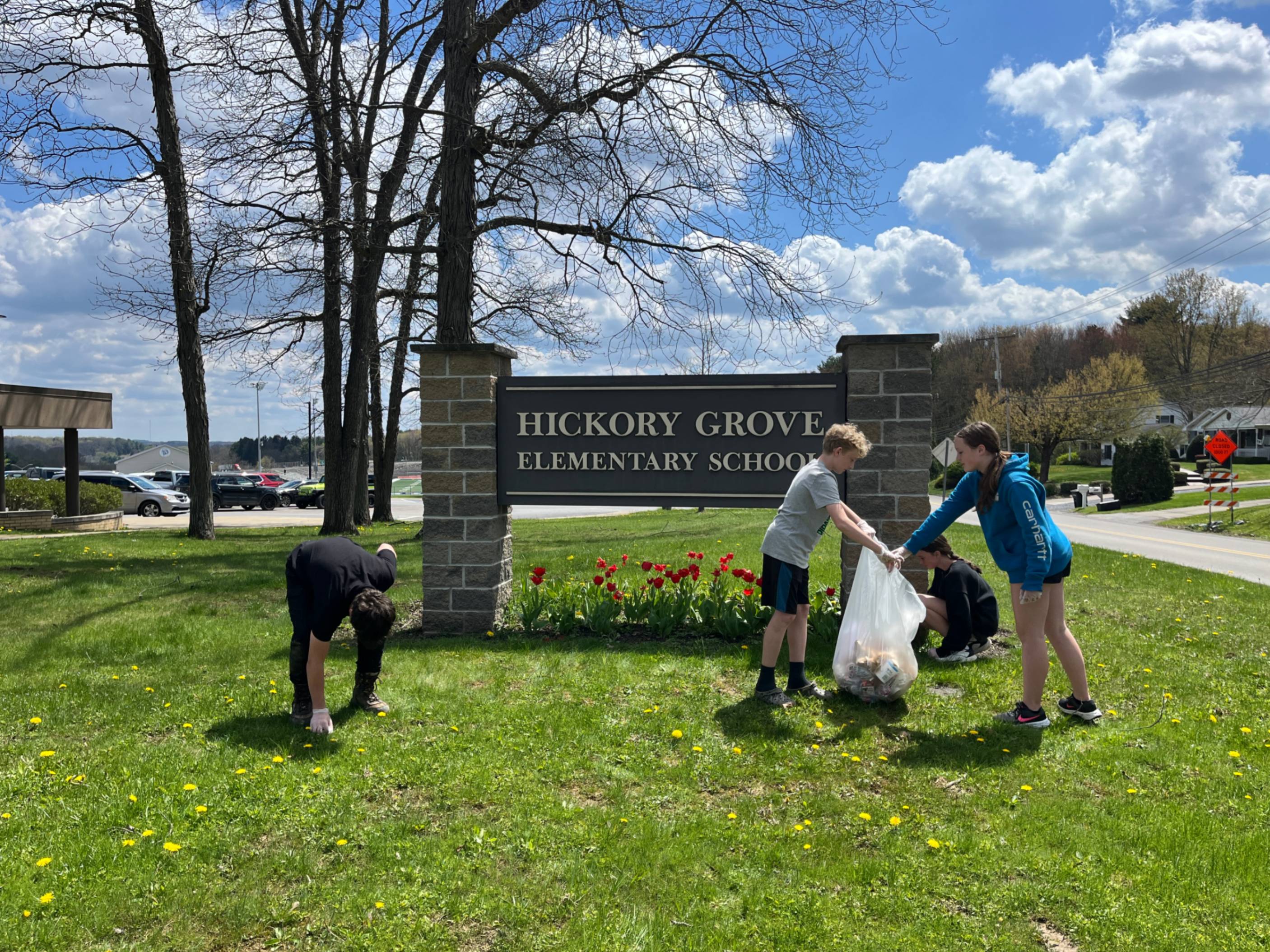 Four Hickory Grove sixth graders cleaning up the grassy area near the school sign.